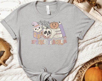 Puede incluir: Camiseta gris claro con un diseño temático de Halloween. El diseño incluye un sombrero de bruja, una telaraña, una calabaza, una calavera, libros y un lápiz. El texto "SPOOKY TEACHER" se muestra en una fuente retro.
