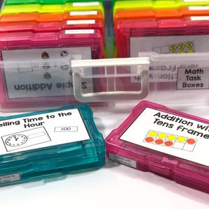 May include: A collection of colorful plastic boxes for educational purposes. The boxes are pink, teal, and clear, with white labels featuring text like "Telling Time to the Hour" and "Addition with Tens Frame." The boxes are stacked and organized.