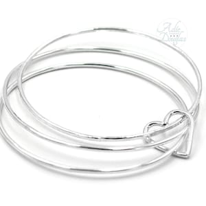 May include: A set of three silver bangles with a heart-shaped charm. The bracelets are thin and round, with a simple, elegant design. The heart charm is attached to one of the bangles.
