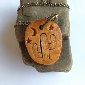 May include: A brown leather necklace with a pendant featuring a carved cactus design with stars and a crescent moon. The necklace is on a brown rock with a gold chain.