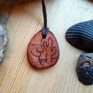 May include: A brown leather necklace with a pendant featuring a hand-drawn image of a face and a cactus. The pendant is a natural stone with a rough texture. The necklace is on a wooden surface.