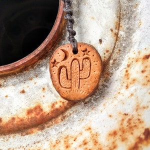 May include: A brown clay pendant with a carved cactus design and two stars. The pendant is hanging from a dark grey cord.