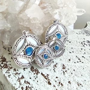 May include: Four silver pendants with a circular design, featuring a central blue gemstone and intricate detailing. The pendants are arranged on a white, textured surface, with a backdrop of clear quartz crystals.