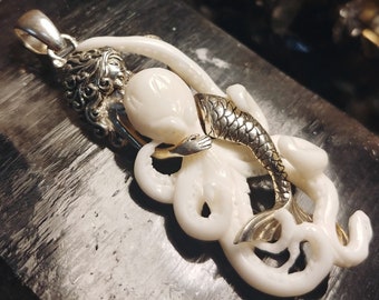 May include: A white and silver pendant featuring a mermaid with a fish tail wrapped around an octopus. The pendant is made of bone and metal.