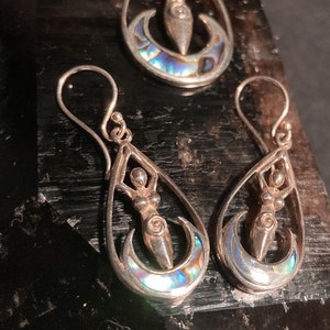 May include: A silver pendant and matching earrings feature a stylized female figure standing on a crescent moon. The pendant and earrings are made of silver and abalone shell.