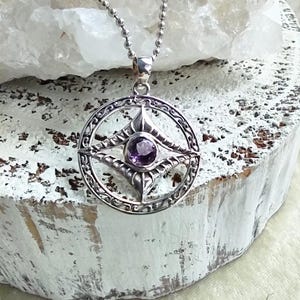 May include: A silver pendant necklace featuring a circular design with an intricate pattern and a central purple gemstone. The pendant hangs from a delicate silver chain. The necklace is displayed on a white, textured surface.