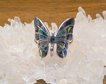 Op de afbeelding: Een vlindervormige ring met een zilverkleurig metalen frame ingelegd met iriserende abalone schelpsegmenten in blauwe, groene en paarse tinten. De vleugels zijn gesegmenteerd met dunne zilveren lijnen en een klein ovaal abalone stukje vormt het lichaam.