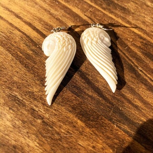 Puede incluir: Un par de pendientes de alas talladas, de color marfil. Cada pendiente presenta un dise&ntilde;o de ala detallado con plumas en capas y una peque&ntilde;a cara en la parte superior. Los pendientes cuelgan de ganchos plateados, perfectos para a&ntilde;adir un toque de elegancia.