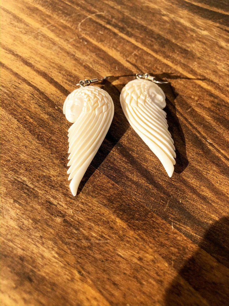 Puede incluir: Un par de pendientes de alas talladas, de color marfil. Cada pendiente presenta un dise&ntilde;o de ala detallado con plumas en capas y una peque&ntilde;a cara en la parte superior. Los pendientes cuelgan de ganchos plateados, perfectos para a&ntilde;adir un toque de elegancia.
