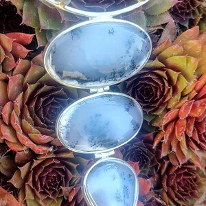 May include: A silver pendant with three teardrop-shaped stones. The stones are a light blue color with a white and gray dendritic pattern. The pendant is hanging against a background of green and red succulents.