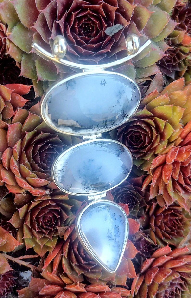 May include: A silver pendant with three teardrop-shaped stones. The stones are a light blue color with a white and gray dendritic pattern. The pendant is hanging against a background of green and red succulents.