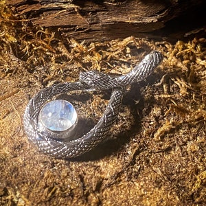 May include: A silver snake pendant with a round moonstone cabochon in the center. The snake is coiled and the moonstone is iridescent.