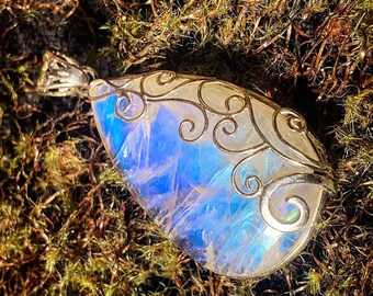 Puede incluir: Un colgante de piedra de luna iridiscente en forma de lágrima con un diseño de filigrana de plata. El colgante está colocado sobre un fondo de musgo verde.