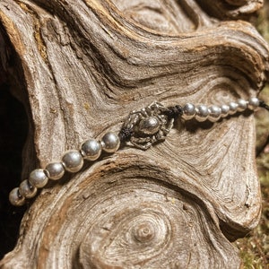 May include: A silver bracelet featuring a spider charm and spherical beads. The bracelet is strung on a black cord and rests on a piece of weathered wood. The spider charm is the focal point of the bracelet.
