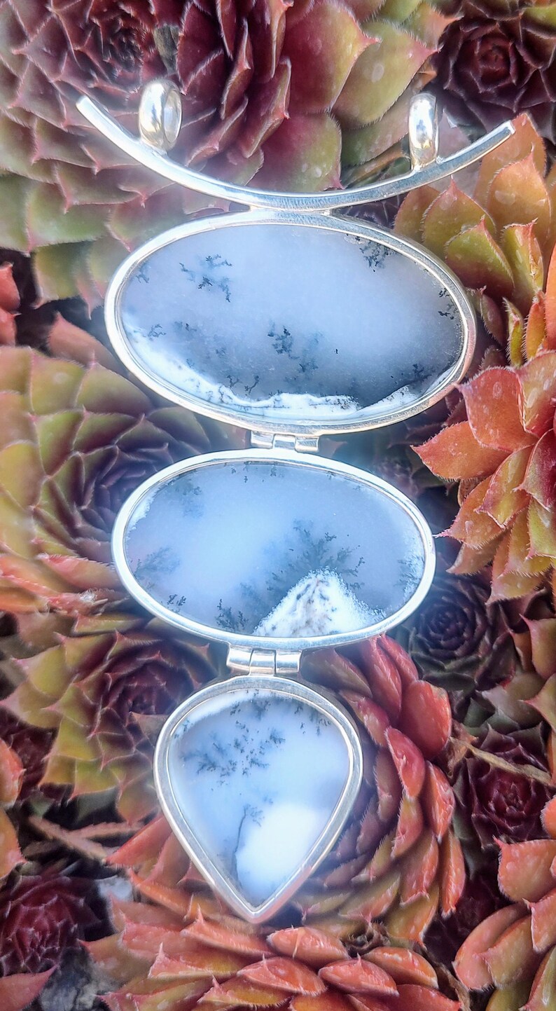 May include: A silver pendant necklace with three oval-shaped agate stones. The stones are a light blue color with dark gray dendritic patterns. The pendant is suspended from a silver chain.