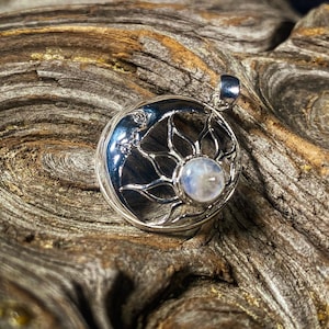 May include: Silver pendant featuring a moonstone set in a sun design. The pendant is on a wooden surface.
