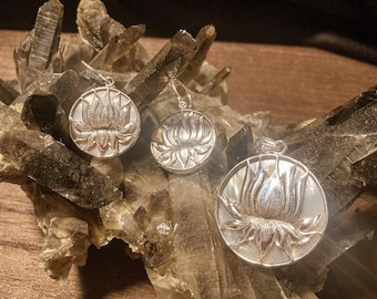 May include: A silver-toned jewelry set featuring a lotus flower design. The set includes a pendant and a pair of earrings, all with a mother-of-pearl background. The lotus flower is intricately detailed, with a polished finish. The jewelry is displayed on a bed of crystals.