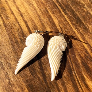 Puede incluir: Pendientes de alas de &aacute;ngel de color marfil con tallado detallado. Cada pendiente presenta una cara esculpida y un dise&ntilde;o de ala en capas, suspendido de ganchos plateados. Los pendientes se muestran sobre una superficie de madera.