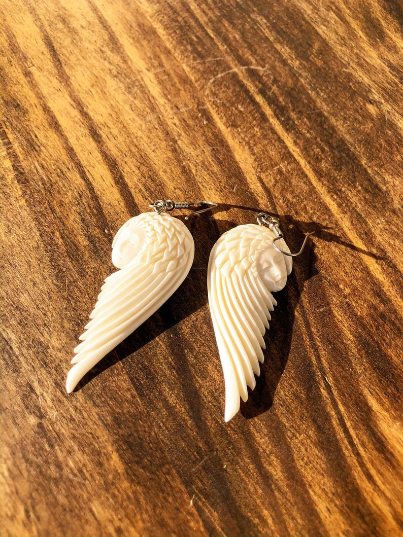 Puede incluir: Pendientes de alas de &aacute;ngel de color marfil con tallado detallado. Cada pendiente presenta una cara esculpida y un dise&ntilde;o de ala en capas, suspendido de ganchos plateados. Los pendientes se muestran sobre una superficie de madera.