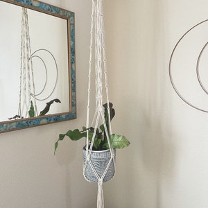Puede incluir: Un macetero de macramé blanco con una maceta de cerámica gris y una planta verde colgando.