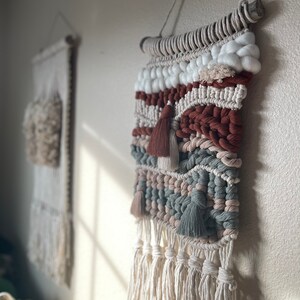 Puede incluir: Un tapiz de pared tejido con una varilla de madera natural en la parte superior. El tapiz presenta una variedad de texturas y colores, incluyendo blanco, marrón, rojo y verde. La parte inferior del tapiz tiene largas borlas blancas.