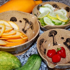Puede incluir: Tres cuencos de cerámica con diseños de hongos, cada uno lleno de diferentes verduras en rodajas. Los cuencos son marrones con un glaseado moteado. Las verduras incluyen calabacín amarillo, pepinos verdes y pimientos rojos.