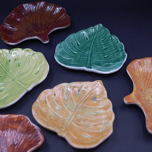 Puede incluir: Un conjunto de seis platos de cerámica con forma de hoja. El set incluye dos platos con forma de ginkgo en rojo oscuro y naranja, y cuatro platos con forma de monstera en varios tonos de verde, verde claro y amarillo, todos con acabado brillante.