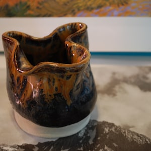 May include: A small, brown and black ceramic pot with a white base. The pot has a unique, wavy design and a glazed finish.