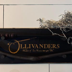 May include: A black wooden sign with the words "Ollivanders Makers of Fine Wands since 382 BC" in gold lettering. Two wooden wands are displayed on the sign.