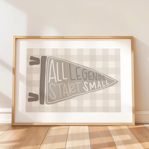 May include: A framed print featuring a pennant design with the text "ALL LEGENDS START SMALL" in a handwritten style. The pennant is gray, set against a light gray and white checkered background. The frame is a light brown color.