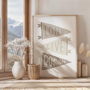 May include: A framed print with three pennant flags, each with text: "LONG", "LIVE", and "BOYHOOD". The flags are in shades of gray and beige. The print is displayed in a room with a window and decorative items.