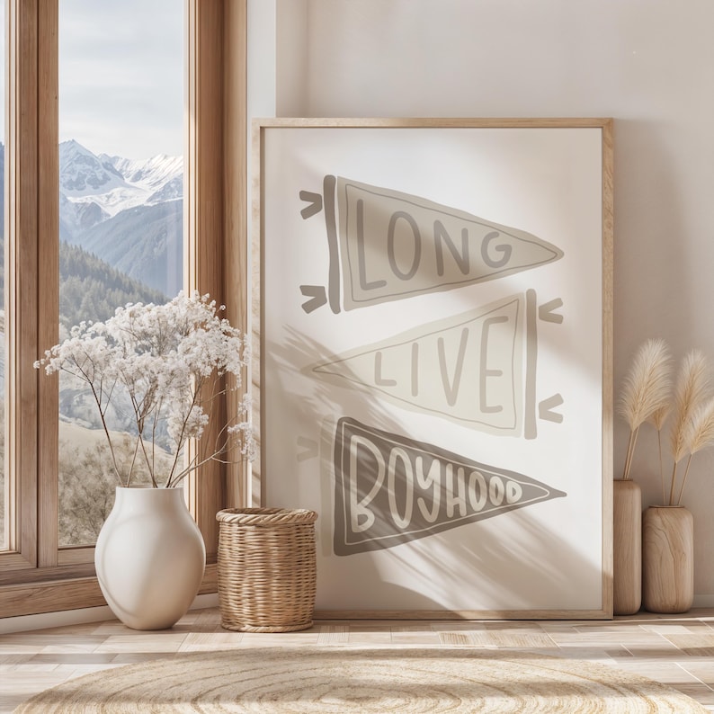 May include: A framed print with three pennant flags, each with text: "LONG", "LIVE", and "BOYHOOD". The flags are in shades of gray and beige. The print is displayed in a room with a window and decorative items.