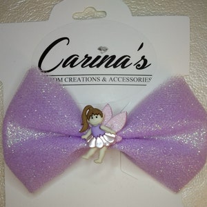 May include: A lavender tulle bow with a sparkly finish and a small fairy figurine in the center. The bow is attached to a white card with the text "Carina's Custom Creations & Accessories".