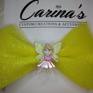 May include: A yellow tulle bow with glitter accents features a small fairy figurine with pink wings and a pink dress. The bow is attached to a white card with the text "Carina's Custom Creations & Accessories".