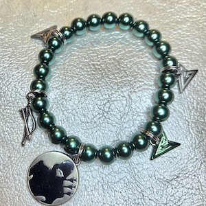 May include: A dark green beaded bracelet with silver-toned charms. The bracelet features round beads and several charms, including arrowheads and a circular pendant with a silhouette design. The bracelet is displayed on a textured, silver surface.