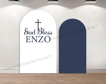 May include: Two arched backdrop panels, one white and one navy blue, stand on a wooden floor. The white panel features the text "God Bless ENZO" with a cross above. The navy panel is a solid color. Overhead, three black pendant lights illuminate the scene.