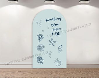 May include: A light blue wedding backdrop with the text "Something Blue Before I Do" in navy blue script. The backdrop features line drawings of hearts, flowers, a starfish, seashells, a cake, and a ring.