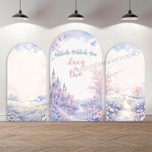 May include: Three arched backdrop covers featuring a watercolour fairy tale scene with a castle, flowers, and a path. The centre panel displays the text Bibbidi-Bobbidi-Boo, Lucy is two in blue and pink script. The design includes soft pastel blue and pink hues.