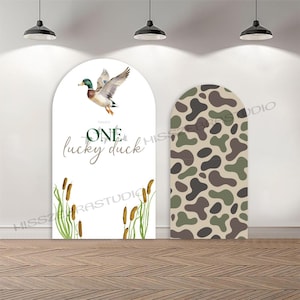 May include: Two large, arched decorative panels. One features a flying duck illustration with the text "One lucky duck." The other panel has a camouflage pattern in shades of green, brown, and tan. The panels are set against a white wall with hanging lights.