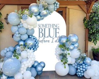 May include: A decorative archway with a white backdrop and the text "SOMETHING BLUE before I do." The arch is adorned with a cascade of blue, silver, and white balloons, accented with floral arrangements. The scene is set outdoors.