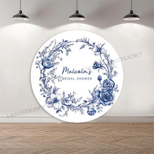 May include: A large, round white backdrop with a blue floral wreath design. The wreath surrounds the text "Malcolms BRIDAL SHOWER" in elegant script. The backdrop is set against a white wall and wooden floor.