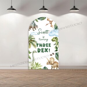 Puede incluir: Un telón de fondo blanco en forma de arco para fiestas con ilustraciones de dinosaurios y plantas tropicales. El texto "Scott is turning THREE REX!" está escrito en verde. Ideal para una fiesta de tercer cumpleaños.