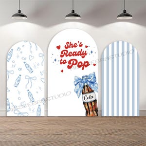 May include: A three-part backdrop for a baby shower. The central panel displays a cola bottle with a blue bow and the text "She's Ready to Pop." The left panel has a blue bottle pattern, and the right panel has blue stripes.