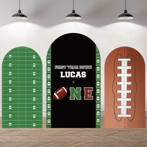 Può includere: Tre sagome di cartone a forma di arco per una festa di primo compleanno. La sagoma centrale è nera con bordo verde e il testo "FIRST YEAR DOWN LUCAS is ONE" con una grafica di un pallone da calcio. La sagoma a sinistra è verde con linee di campo e numeri bianchi. La sagoma a destra è marrone con cuciture e lacci bianchi.