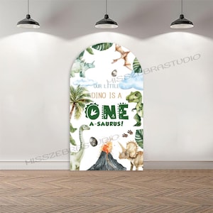Op de afbeelding: Een witte boogvormige achtergrond met een dinosaurus thema. Het bevat illustraties van verschillende dinosaurussen, palmbomen en een vulkaan. De tekst "OUR LITTLE DINO IS A ONE A-SAURUS!" is prominent aanwezig.
