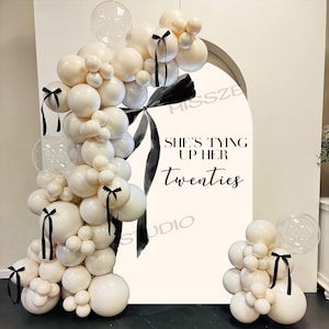 Puede incluir: Un arco de globos blancos y crema, acentuado con cintas negras. Un gran letrero con el texto "SHE'S TYING UP HER Twenties" está en el fondo. También se incluyen globos transparentes en forma de burbuja.