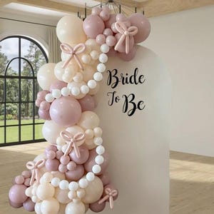 May include: A decorative balloon arch in cream and pink hues, featuring bows and a string of white beads. The words "Bride To Be" are displayed on a white backdrop, suitable for a bridal event or wedding.