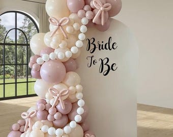 May include: A balloon arch in shades of pink and cream, adorned with decorative bows and a string of white beads. The words "Bride To Be" are written on a white backdrop, perfect for a bridal shower or wedding celebration.