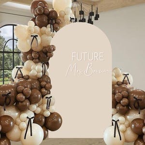 Può includere: Un arco di palloncini nei toni del marrone e crema, adornato con nastri neri. L'arco incornicia uno sfondo beige con la scritta "FUTURE Mrs. Brown" in bianco, ideale per un addio al nubilato o un matrimonio.