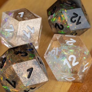 以下が含まれることがあります： キラキラと輝く、12面体のサイコロが4つ。2つは黒い数字の金色のグリッター入り、2つは白い数字のクリアグリッター入り。サイコロは木製の背景に置かれています。テーブルゲーム用のサイコロです。